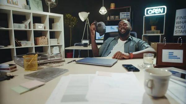 Cheerful african american businessman fanning face with cap in late coworking.