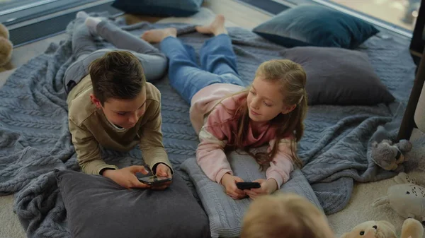 Kinderen die smartphones spelen. broers en zussen liggend met mobieltjes volgende kind op tapijt. — Stockfoto