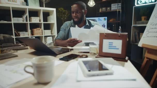 Gericht afro man controleren van gegevens van laptop scherm met documenten binnen. — Stockvideo