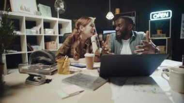 Gülümseyen afro adam karanlık ofisteki dizüstü bilgisayarın yanındaki hipster kadına hikaye anlatıyor..