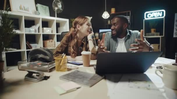 Sorrindo afro homem contando história para mulher hipster perto de laptop no escritório escuro. — Vídeo de Stock
