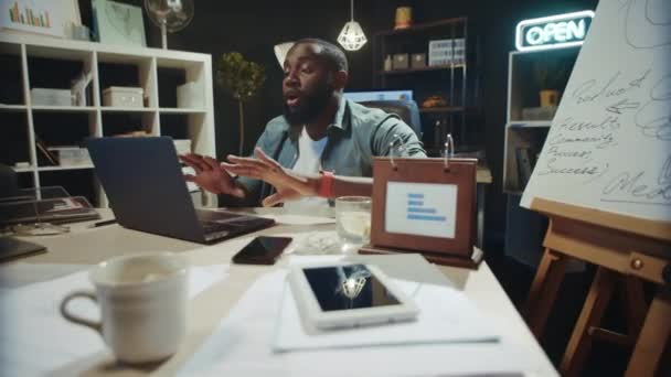 Emotionaler afrikanischer Mann, der im dunklen Büro online spricht. Afro kerl suche kamera — Stockvideo