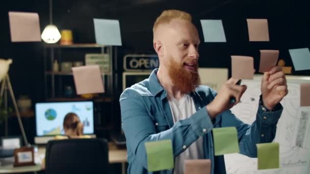 Крупный план улыбающийся хипстер приклеивает заметки на стеклянную доску в ночном офисе — стоковое видео