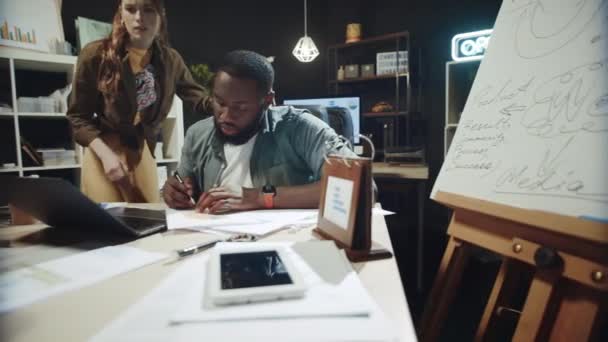 Kvinnlig professionell kontroll arbete afro man nära laptop i mörkt kontor. — Stockvideo