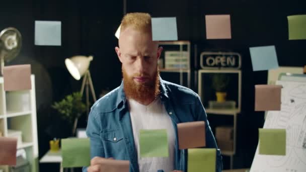 Šťastný podnikatel lepení barevné papíry na skleněnou tabuli v hipster kanceláři. — Stock video