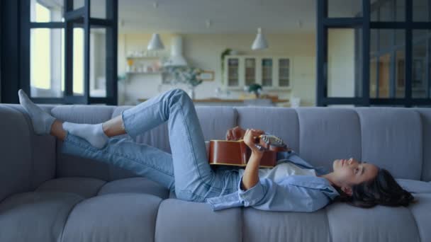 Chica tocando la guitarra en el salón. Mujer tumbada en un sofá con instrumento musical — Vídeo de stock