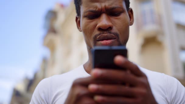 Un afro frustrado usando el móvil en la calle. Hombre mensaje de texto en el teléfono inteligente — Vídeos de Stock