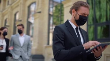 Açık havada tablet kullanan maskeli bir işadamı. Takım elbiseli adam sosyal mesafeyi koruyor.