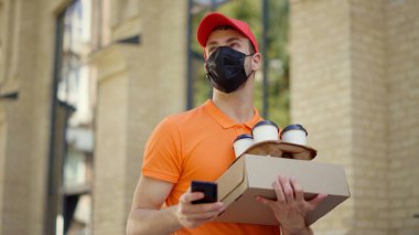 Maskeli kurye dışarıda akıllı telefon kullanıyor. Siparişleri taşıyan teslimatçı..