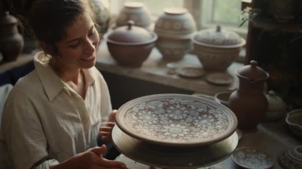 Glückliches Mädchen untersucht Teller in Werkstatt. Keramiker spielt mit Töpferscheibe — Stockvideo