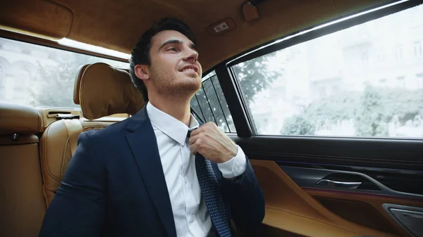 Relaxed male ceo sitting in comfortable car afterwork. Business man fixing tie