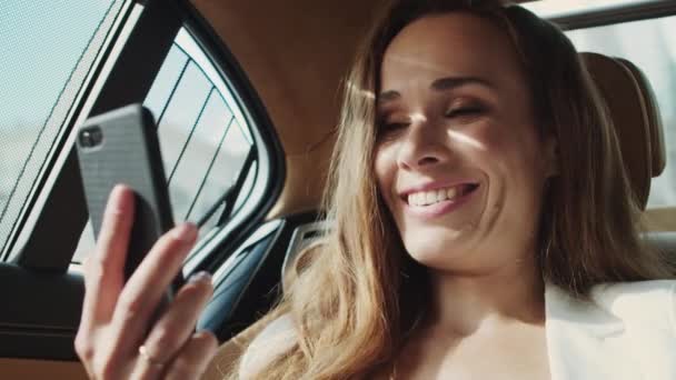 Vriendelijke zakenvrouw glimlachend op video bellen op smartphone in luxe moderne auto. — Stockvideo