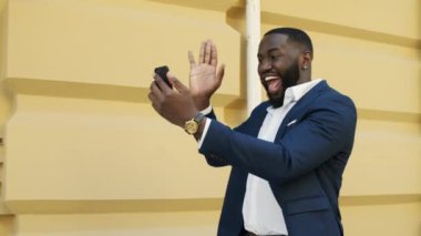 Afro iş adamı dışarıda video görüşmesi yapıyor. Afro adam telefonla el kol hareketi yapıyor.