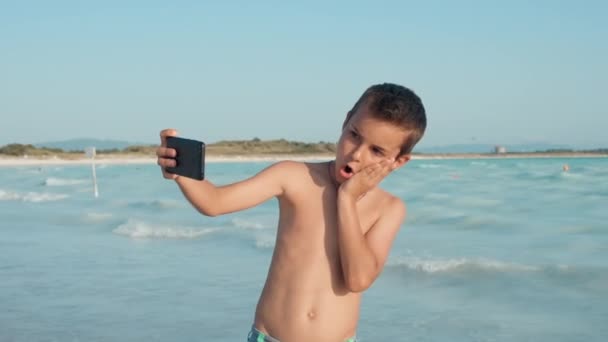 En leende kille njuter av sommarsemestern vid havet. Lycklig pojke grimaserar på stranden. — Stockvideo
