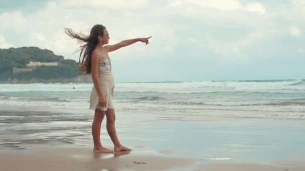 Chica despreocupada mirando al horizonte en la playa. Adolescente apuntando al cielo a la orilla del mar. — Vídeo de stock