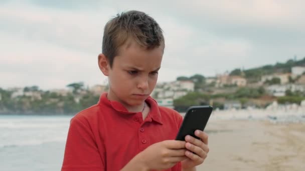 Fokuserad pojke tillbringar tid på stranden. Seriös kille som letar efter mobiltelefon vid havet. — Stockvideo