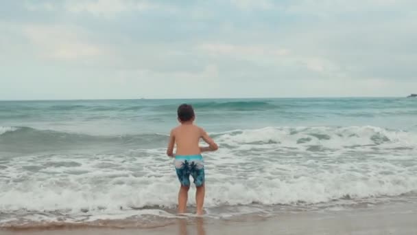 Szczęśliwy chłopiec cieszący się latem na plaży. Wesoły chłopak skaczący w falach nad morzem. — Wideo stockowe