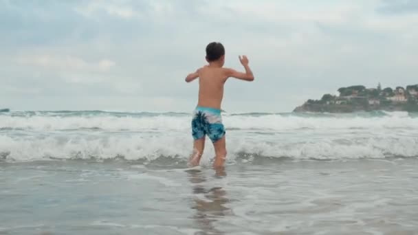 Szczęśliwy chłopiec bawiący się na wybrzeżu. Beztroski facet tańczący w wodzie na brzegu morza. — Wideo stockowe