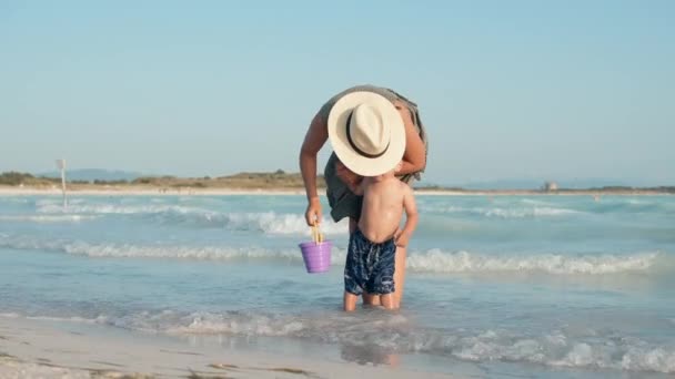 Молодая мать разговаривает с сыном на берегу моря. Милый мальчик, идущий вдоль серфинга. — стоковое видео