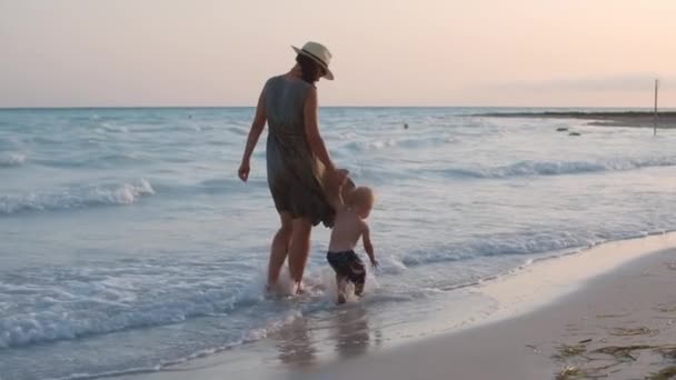 Netter kleiner Junge sitzt im Wasser am Meer. Mutter und Sohn spazieren am Strand. — Stockvideo