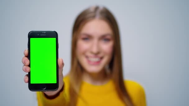 Mujer sonriente mostrando teléfono inteligente con pantalla verde en el estudio. Chica pulgares hacia arriba — Vídeos de Stock