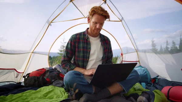 Guy sitting in camping tent with laptop . Ginger man using touchpad on laptop