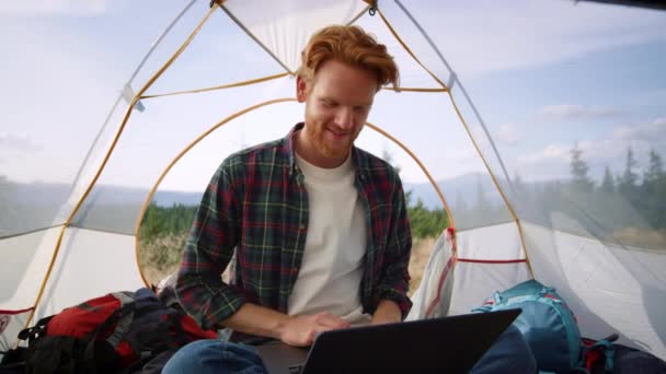 Рыжий парень использует ноутбук во время похода. Мужчина фрилансер, работающий на ноутбуке в палатке — стоковое видео