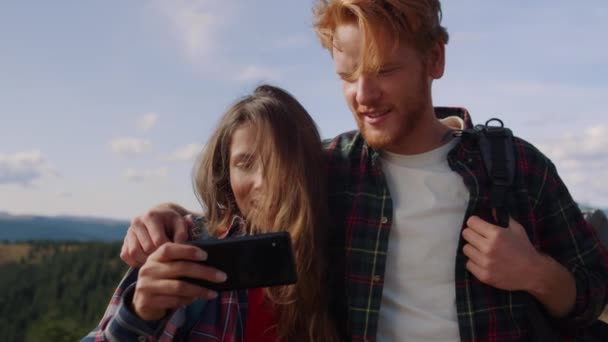 Gülümseyen adam ve kız dağlarda yürüyüş yaparken akıllı telefondan video izliyorlar. — Stok video