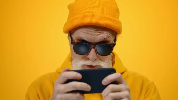 Gammal man som letar efter mobiltelefon i studio. Fokuserad kille spelar online inomhus. — Stockfoto