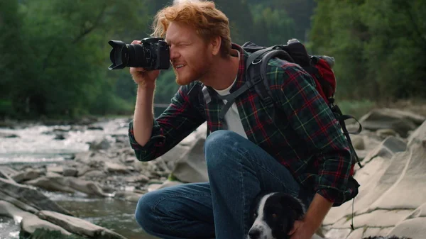 Φωτογράφος κάθεται στην όχθη του ποταμού με το σκυλί. Ο τύπος τραβάει φωτογραφίες στην κάμερα. — Φωτογραφία Αρχείου