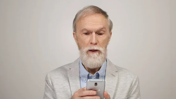 Vreugdevolle man die binnenshuis nieuws leest over de telefoon. Oude man met mobiele telefoon in de studio. — Stockfoto