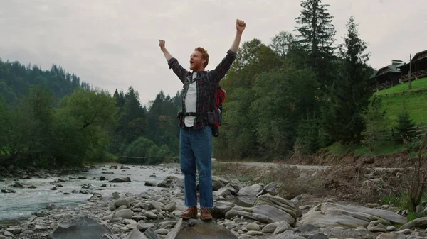 Άνδρας πεζοπόρος στέκεται στη βραχώδη όχθη του ποταμού στα βουνά. Κοκκινομάλλης σηκώνει τα χέρια. — Φωτογραφία Αρχείου