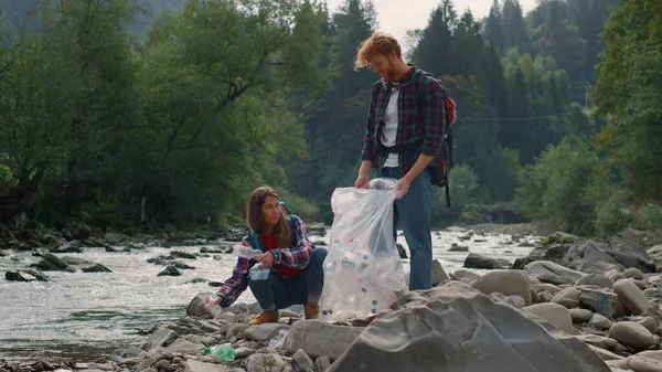 Πεζοπόροι καθαρίζουν την όχθη του ποταμού από πλαστικό. Χαρούμενος άντρας και γυναίκα μαζεύουν σκουπίδια. — Φωτογραφία Αρχείου