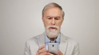 Odaklanmış yaşlı adam internet haberlerini cep telefonuyla tarıyor. Stüdyodaki gri duvarın yanında..