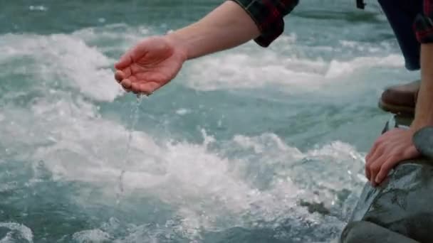 Un tipo sentado en la orilla rocosa del río. Caminante tocando agua con la mano en el flujo del río — Vídeo de stock