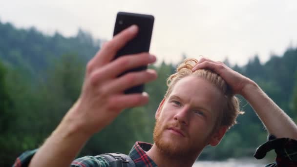 Мужчина чинит прическу перед селфи. Парень делает селфи на смартфоне в горах — стоковое видео
