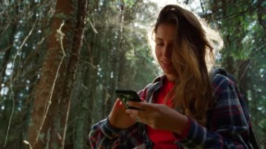 Yaz ormanında akıllı telefon kullanan bir kadın. Gülümseyen yürüyüşçü cep telefonunu kaydırıyor