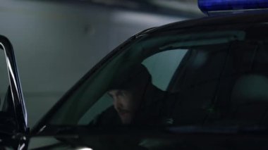 Polis memuru polis arabasının kapısında duruyor. Polis silahı polis aracına doğrultuyor.