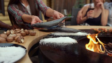 Bilinmeyen kız ızgaraya tuz basıyor. Kadın dışarıda balık hazırlıyor.