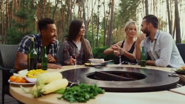 Młodzi ludzie rozmawiają o przygotowaniach żywności na zewnątrz. Przyjaciele siedzą przy grillu — Wideo stockowe
