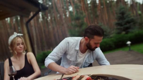 Gente positiva sentada alrededor del fuego afuera. Chico lanzando leña en barbacoa parrilla — Vídeos de Stock