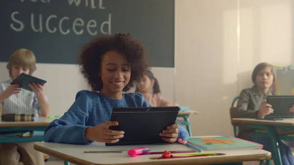 Glimlachen Afrikaans amerikaans meisje met behulp van digitale tablet in de klas met klasgenoten — Stockfoto