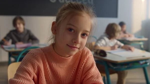 Cute pupil sitting at desk at elementary school. Smiling girl looking at camera