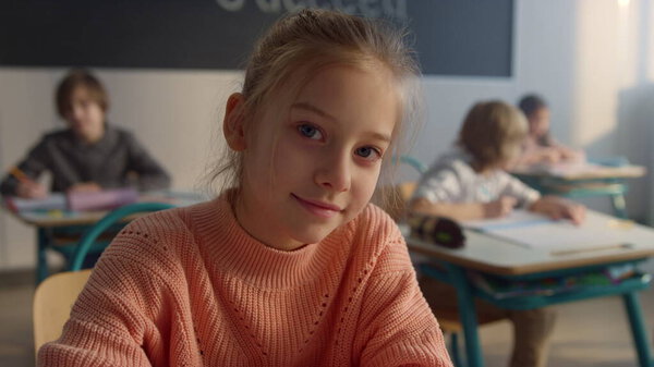 Cute pupil sitting at desk at elementary school. Smiling girl looking at camera 