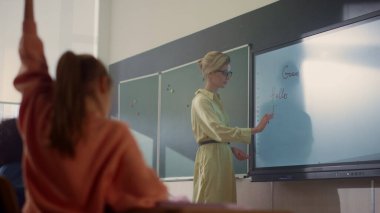 Öğretmen interaktif dijital yazı tahtasında yazıyor. Öğrenciler el kaldırıyor.