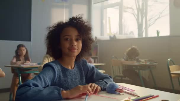 Meisje kijkt naar de camera in de klas. Vrolijke student zit aan het bureau op school — Stockvideo