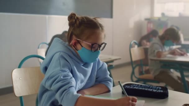 Meisje in het gezicht masker leren in de klas. Schoolmeisje schrijven in notebook — Stockvideo