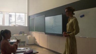 Bayan öğretmen çocuklarla dolu sınıfın önünde tahtada duruyor.