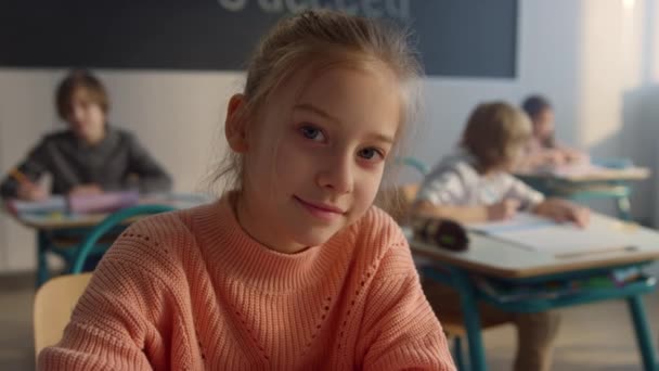 Leuke leerling aan het bureau op de basisschool. Glimlachend meisje kijkend naar camera — Stockvideo