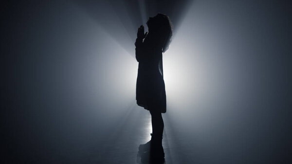 Silhouette of beautiful woman praying in floodlight background. Side view young female person saying prayer in darkness. Full length of religious girl folding hands for prayer indoors.